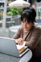 A woman is working remotely at a cafe, sitting outdoor, using her laptop and reading a book. photo
