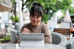 un feliz, positivo asiático mujer es trabajando de forma remota, utilizando su digital tableta a un al aire libre salón. foto