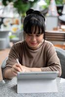 un contento asiático mujer trabajando remotamente desde un café en el ciudad, utilizando su digital tableta. foto