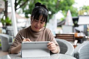 un contento asiático mujer trabajando remotamente desde un café en el ciudad, utilizando su digital tableta. foto