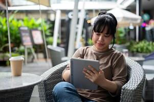 un feliz, positivo asiático mujer es trabajando de forma remota, utilizando su digital tableta a un al aire libre salón. foto