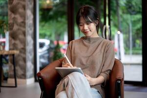 un hermosa asiático mujer es escritura en su tableta con un aguja bolígrafo, trabajando remotamente desde un cafetería. foto