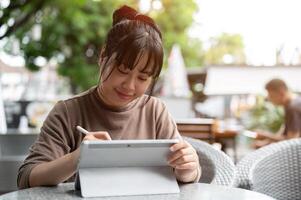 un contento asiático mujer trabajando remotamente desde un café en el ciudad, utilizando su digital tableta. foto