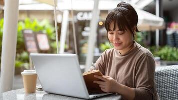 un mujer es trabajando remotamente a un cafetería, sentado exterior, utilizando su ordenador portátil y leyendo un libro. foto