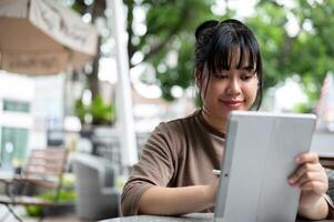 un contento asiático mujer trabajando remotamente desde un café en el ciudad, utilizando su digital tableta. foto