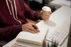 Cropped image of a man sitting in front of a computer, taking notes in a book, working in a room. photo