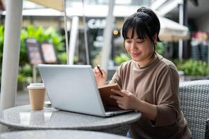un mujer es trabajando remotamente a un cafetería, sentado exterior, utilizando su ordenador portátil y leyendo un libro. foto