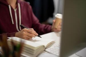 Cropped image of a man sitting in front of a computer, taking notes in a book, working in a room. photo