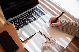 A top-view image of a woman is taking notes in her notebook while working on her laptop indoors. photo