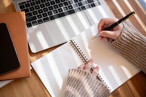 A top-view image of a woman is taking notes in her notebook while working on her laptop indoors. photo