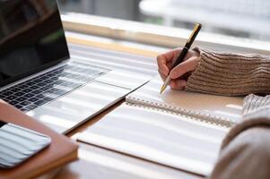 Close-up image of a woman is writing something in her notebook and working on her laptop indoors. photo