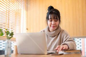 A positive Asian woman in a cosy sweater is working on her laptop computer in a cafe or library. photo