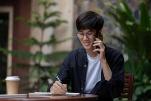 A happy Asian man is taking notes in his book while talking on the phone with someone outdoors. photo