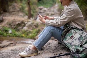 A female trekker is sitting on a rock and using her phone, taking a break while hiking. photo