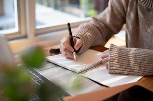 Close-up image of a woman is writing something in her notebook and working on her laptop indoors. photo