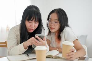 Two Asian female friends are watching something on a smartphone together while sitting in a cafe. photo