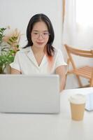 A young Asian woman wearing eyeglasses is working remotely from a cafe, working on her laptop. photo