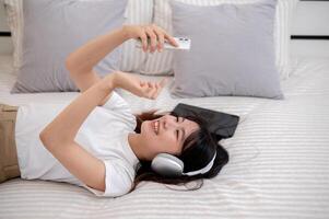 A woman is lying on the bed, using her smartphone, and enjoying listening to music on her headphones photo