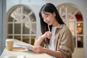 un joven hermosa asiático mujer en casual vestir es sentado en un café y utilizando su digital tableta. foto