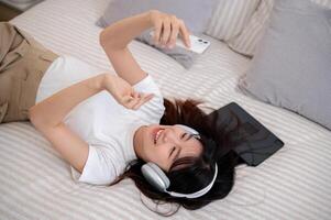 A woman is lying on the bed, using her smartphone, and enjoying listening to music on her headphones photo
