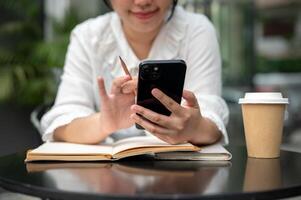 A woman using her smartphone while writing something in her notebook at a table. photo