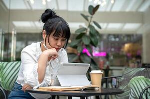 un joven asiático mujer leyendo alguna cosa en su digital tableta, trabajando remotamente desde un café tienda. foto