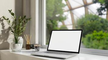 A laptop computer white-screen mockup on a white table against the window with a green nature view. photo