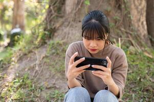 Young Asian woman focuses on a game on her smartphone while sitting under a tree in a park. photo