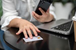 A woman is touching a credit card on a table, looking at her credit card number and using her phone. photo