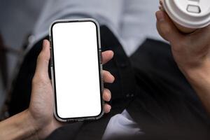 Close-up image of a man holding a white-screen smartphone and a coffee cup while sitting outdoors. photo