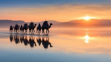 Silhouette of a camel caravan walking along a beach at sunset, with reflection on water photo