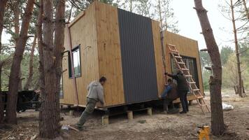 Wooden Prefabricated Cabin Installation in Forest Setting. Three men are seen working on the cabin as it is being placed on its foundation. The cabin has a modern design with a wooden exterior video