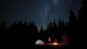 A man with a lantern near a bonfire and tent in night forest. The Stars and Milky Way move in the night sky. Travel concept. 4K video