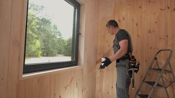 A man applies acrylic paint to a wall in a cabin with a large window. The cabin has a natural wood interior and is located in a forested area. video