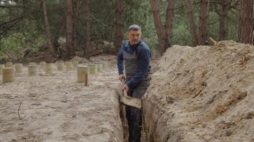 A worker wearing is actively digging a narrow trench in a forested area. The surrounding terrain is uneven with visible tree stumps and soil heaps. video