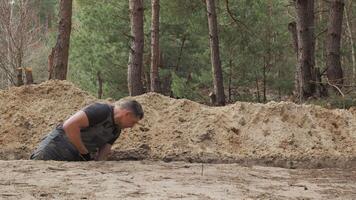 A worker wearing is actively digging a narrow trench in a forested area. The surrounding terrain is uneven with visible tree stumps and soil heaps. video
