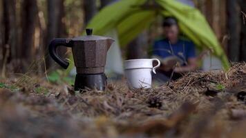 A coffee pot and a mug in the foreground in the forest. A man reads a book in a tent on the background. Travel and healthy lifestyle concept video