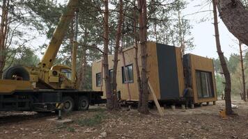 Wooden Prefabricated Cabin Installation in Forest Setting. Two men are seen working on the cabin as it is being placed on its foundation. The cabin has a modern design with a wooden exterior video