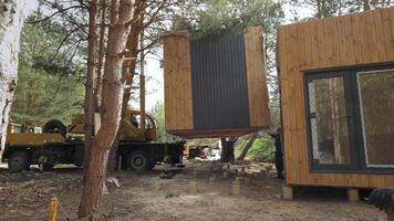 A crane lifts a prefabricated cabin into place in a wooded area. The cabin is made of wood and has a modern design. Process of constructing a small, off-grid home in a remote location. video