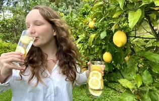 it's just a lemon paradise, a teenage girl stands against the backdrop of a lemon tree with lemons and drinks lemonade from a glass glass in her other hand, she holds a jug of drink photo