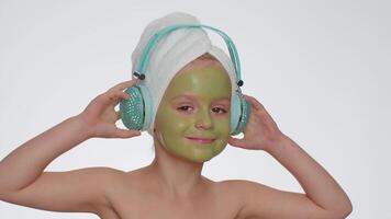 Smiling young child girl moisturizing green mask on face listening to music on headphones, dancing video