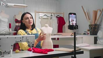 Seamstress runs an online blog where she explains how to sew accurately on a white mannequin in her studio. She records a using her phone, which is mounted on a tripod. Medium shot. Slow motion. video