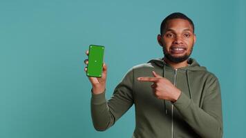 Portrait of african american man doing influencer marketing using green screen phone, studio background. BIPOC person holding empty copy space mockup smartphone device, camera A video