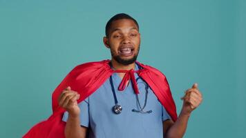 Male nurse portraying superhero showing courage and strength fighting diseases, studio backdrop. Portrait of african american doctor posing as hero in costume, holding empowering speech, camera A video