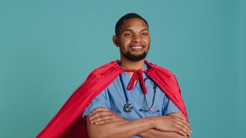 Smiling male nurse portraying superhero wearing red cape, isolated over studio background. Portrait of cheerful heroic medic posing as hero in costume with mantle, camera A video