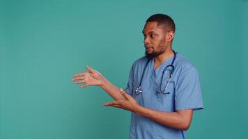 Portrait of african american nurse pointing hands towards empty space, doing advertising, talking with audience. Healthcare employee doing promotional ad, showing copy text, studio backdrop, camera B video