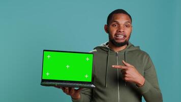 Smiling man holding isolated screen laptop, doing recommendation, talking with audience. Cheerful person pointing towards chroma key notebook, studio background, camera A video