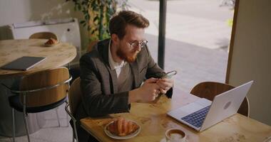 Businessman Reading News In Smartphone Sitting In Cafe, Open Laptop On Table, Using Mobile App video