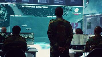 Staff sergeant monitoring satellite live imagery on a big screen, gathering information for future military operations. Army officer employs reconnaissance system in command post. Camera A. video