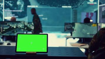 Tablet with greenscreen display running in a command post base of operations, military personnel ensuring national safety in control room. Soldiers and commanders use radar surveillance. Camera B. video
